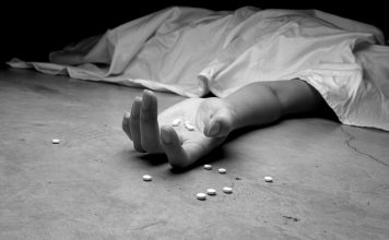 A black and white image of an addict, dead beneath a sheet, arm outstretched, pills in the palm of their lifeless hand and littering the floor near their hand.