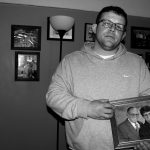 A man, David Stanley, stands holding his parents photo, behind him the wall is filled with other family photos. .