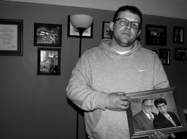 A man, David Stanley, stands holding his parents photo, behind him the wall is filled with other family photos. .