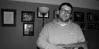 A man, David Stanley, stands holding his parents photo, behind him the wall is filled with other family photos. .