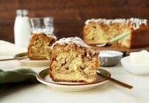 Cinnamon Roll Coffee Cake A slice of Cinnamon Roll Coffee Cake sits on a plate in the foreground with the remainder of the cake in the background.