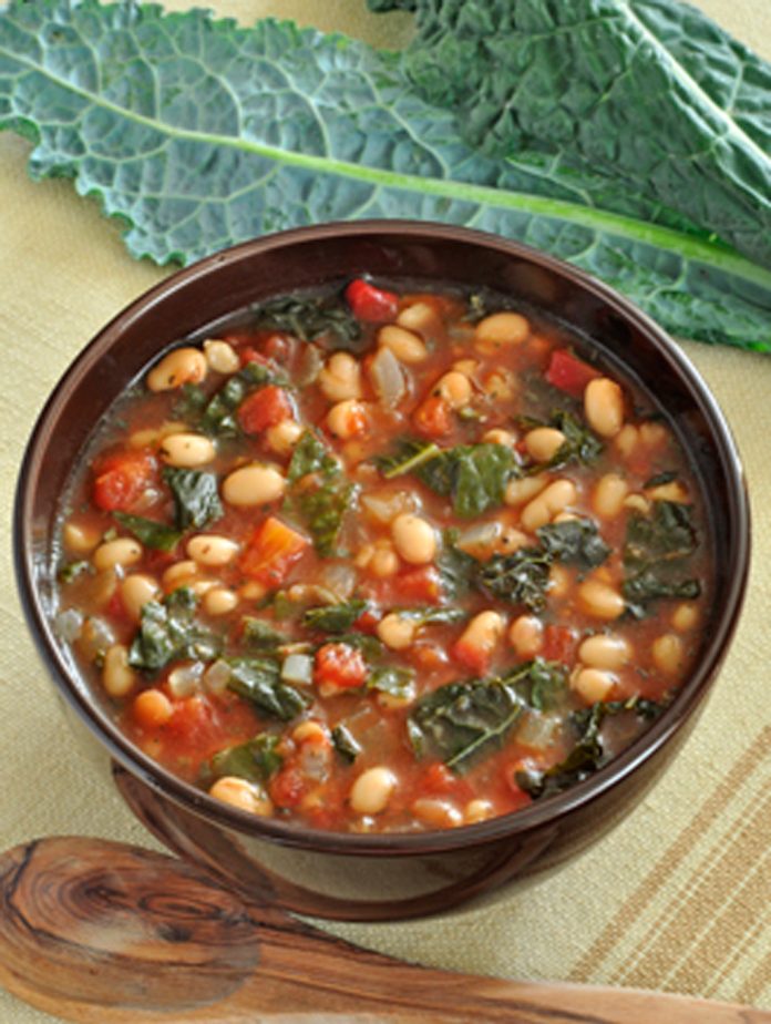 A bowl of kale and white bean soup with Kale leaves in the background.