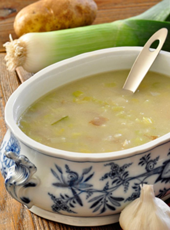 A bowl of Creamy Potato Leek Soup, a leek and poato are in the background and a bulb of garlic is in the foreground