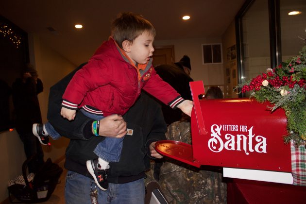 A boy puts a letter for Santa into a mailbox while his father holds him up so he can reach.
