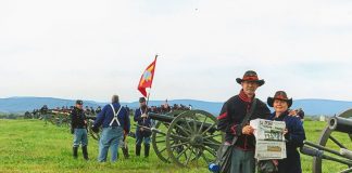 The Dazeys stand dressed in Union soldier uniforms on the battle field. A group of Union soldiers stand behind them near an Civil War era cannon.