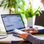 Ohio moves forward again on broadband expansion a person sits at a computer, holding a phone in one hand, with glasses on the desk next to her.