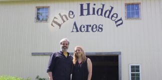 A man and a woman stand in front of a barn with the words "The Hidden Acres" painted on it.