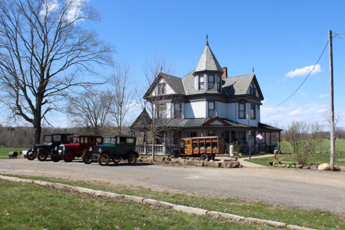 antique cars sit outside a victorian home. A home built hearse sits in front of the home with a casket in the back.