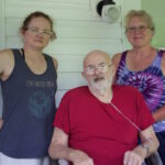 Two women and a man on the porch of a farmhouse.