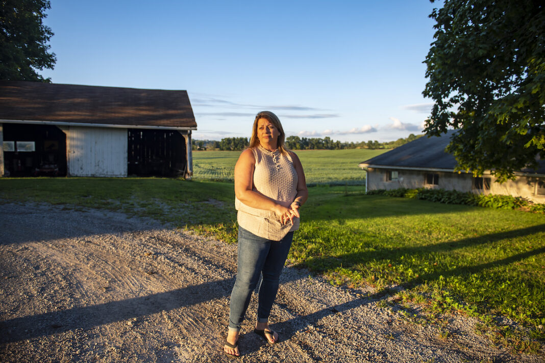 A woman wearing jeans and a light-colored top stands on a gravel driveway, framed in the background by a small open barn and a tree. Behind her are soybeans in a field.