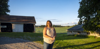 Balancing love, loss and life after a farmer’s suicide A woman wearing jeans and a light-colored top stands on a gravel driveway, framed in the background by a small open barn and a tree. Behind her are soybeans in a field.