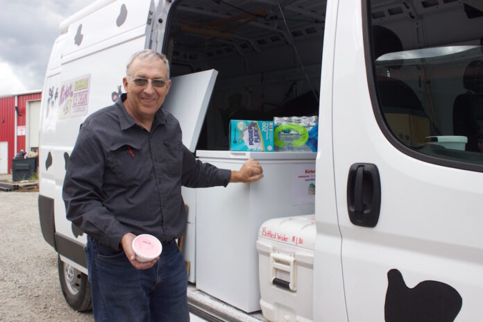 porterfield_farms_and_ice_cream_5 A man holds a cup of ice cream next to a van with freezers inside it and a cow pattern painted on the outside.