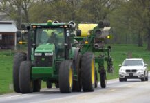 Farmers ask drivers to be patient during spring planting season A tractor pulling a planter drives on a road with a car following closely behind it.
