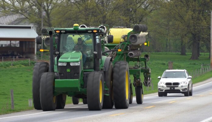 tractor on a road A tractor pulling a planter drives on a road with a car following closely behind it.