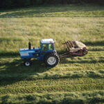 a blue tractor moves across a field of green, mowing hay