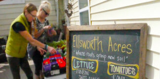 Two women stand next to table full of plants with sign in the foreground that says Ellsworth Acres