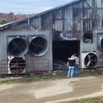 Two women with their backs to the camera wrap their arms around each other as they look at a damaged and burned dairy barn.