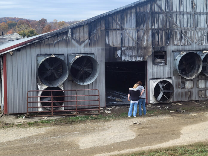 Two women with their backs to the camera wrap their arms around each other as they look at a damaged and burned dairy barn.