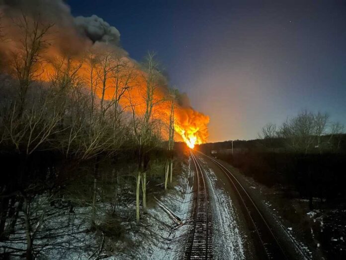 train derailment photo A flame reaching high into the sky can be seen at the end of a railroad tracks that go up the middle of the photo.